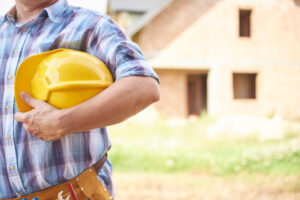 Construction worker in a plaid shirt holds a yellow hard hat at a building site.
