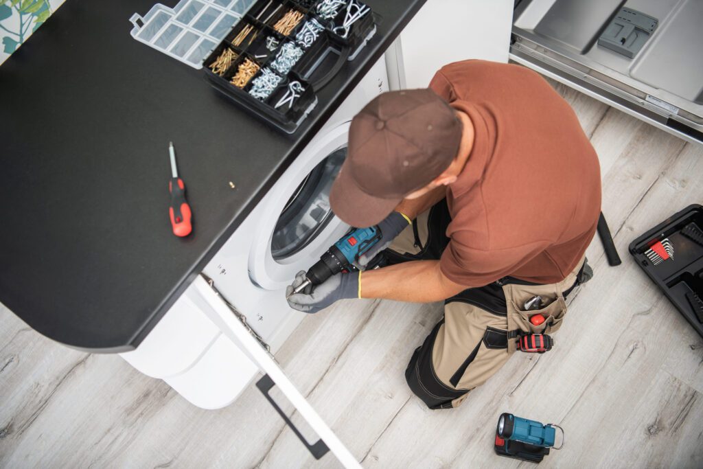 Repair technician kneels beside a front-loading washing machine, using a cordless drill with a tool box and small parts nearby on the counter.
