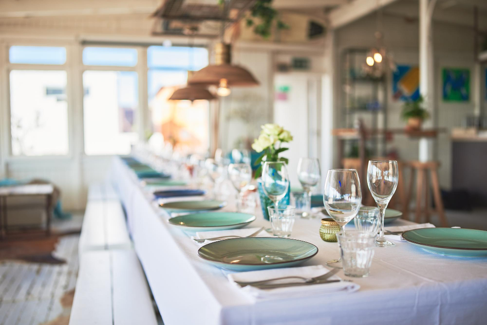 Long dining table set for a meal with green plates, clear glassware, and flowers in a bright room.