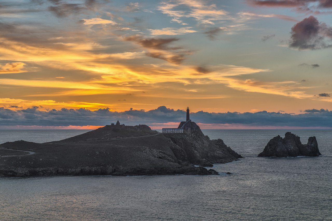 Faro Vilan - A Coruna - IP - Noroeste Noticias 24h Lighthouse on a rocky island at sunset, with a warm orange sky and calm sea surrounding it.